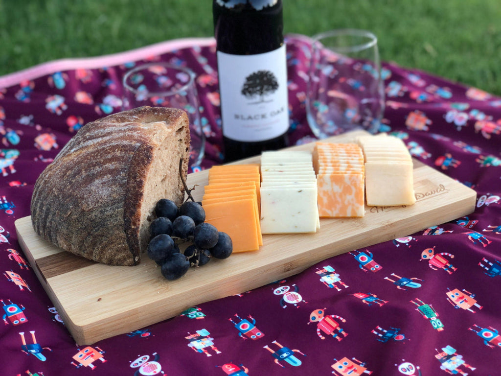 Wooden board with bread, cheese, and grapes on a patterned blanket outdoors